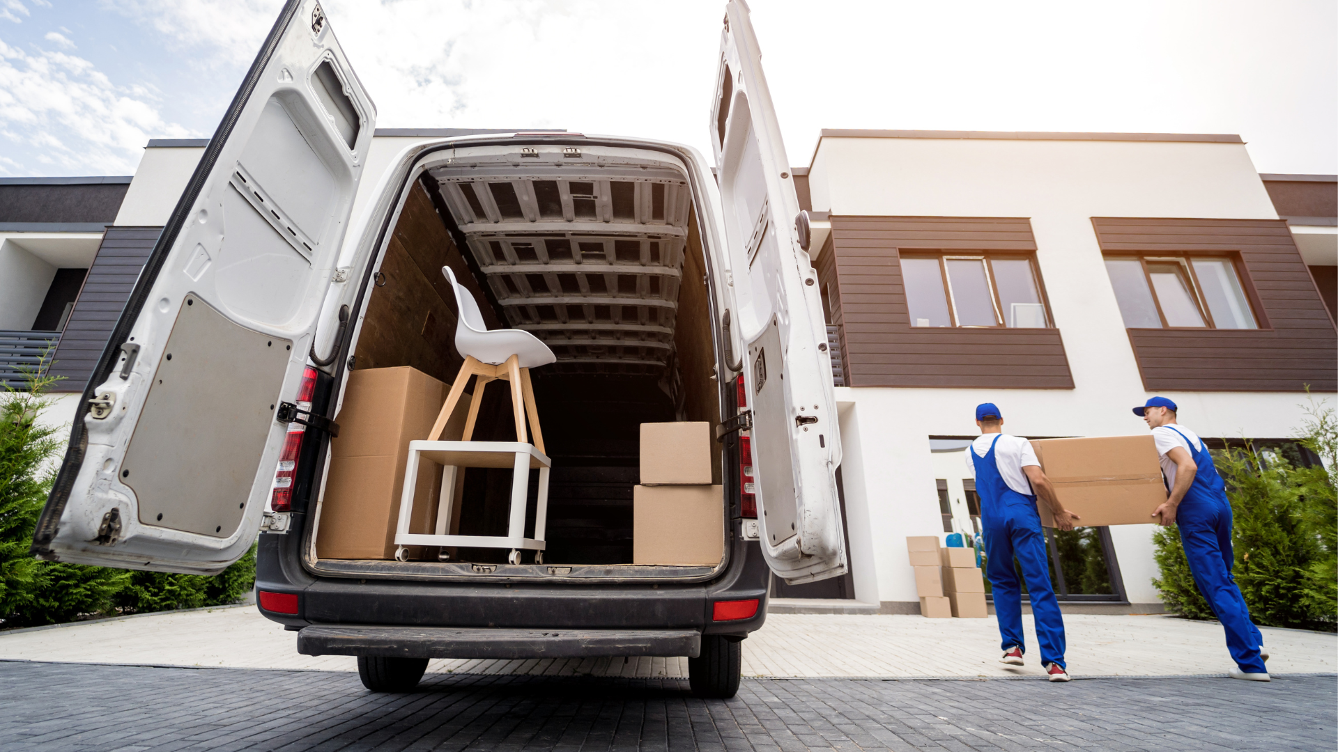 Two movers in blue uniforms carry a large cardboard box toward a modern apartment building, with the back of a moving van open and filled with boxes and furniture showcasing the services of a trusted Jersey City moving company.