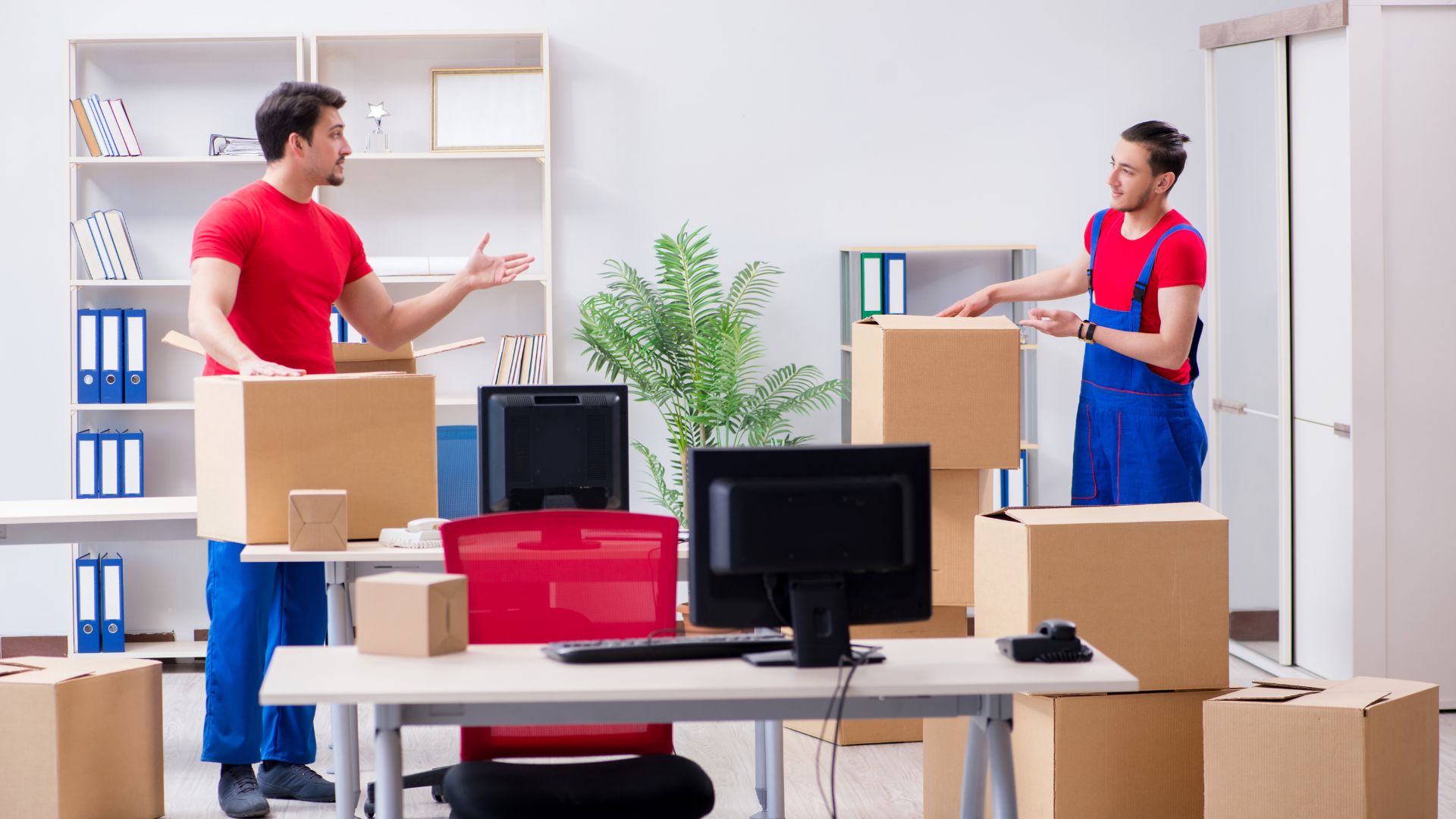 Two professional movers in red shirts and blue overalls packing cardboard boxes in an office space, with desks, computers, and shelves in the background. Keyword: movers Newark NJ.