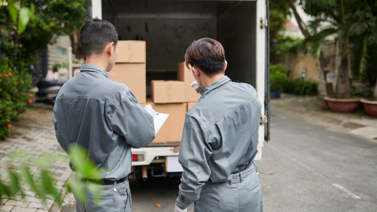 Two workers from a Jersey City moving company wearing gray uniforms are standing at the back of a truck, checking and loading cardboard boxes for delivery.